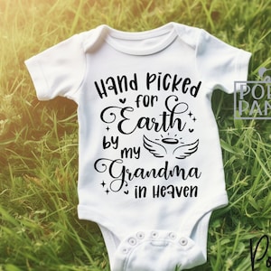 May include: White baby onesie with black text that reads "Hand Picked for Earth by my Grandma in Heaven". The onesie is lying on a bed of green grass.