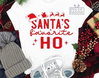 May include: White t-shirt with a red graphic that reads "SANTA'S favorite HO" with Santa and reindeer. The shirt is paired with a gray beanie, jeans, and gray sneakers. A Christmas-themed design.