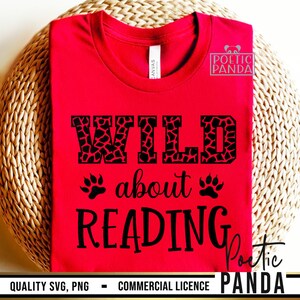 Puede incluir: Una camiseta roja con las palabras "WILD about READING" en negro. La palabra "WILD" tiene un estampado de jirafa. Huellas de patas est&aacute;n a ambos lados de la palabra "about". La camiseta est&aacute; sobre una superficie tejida de color marr&oacute;n claro.
