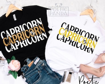 Op de afbeelding: Twee T-shirts met het woord "Capricorn" drie keer herhaald in zwart en gouden glitter. Het woord "Capricorn" is op elkaar gestapeld, met het bovenste woord in zwart, het middelste woord in gouden glitter en het onderste woord in zwart. De T-shirts hangen aan een houten hanger en liggen op een blauwe denim achtergrond.