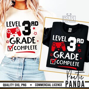 May include: A white and black t-shirt with the text "Level 3rd Grade Complete" and a red and black video game controller graphic. The text is in red and black. The t-shirt is on a person wearing blue jeans.