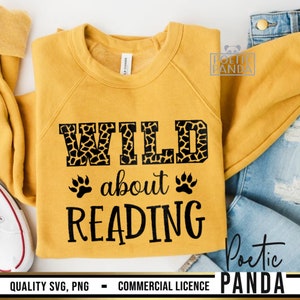 Puede incluir: Sudadera amarilla mostaza con el texto negro "WILD about READING". La palabra "WILD" tiene un estampado de jirafa. Huellas de patas a ambos lados de la palabra "about". La sudadera est&aacute; doblada sobre una superficie blanca con vaqueros.