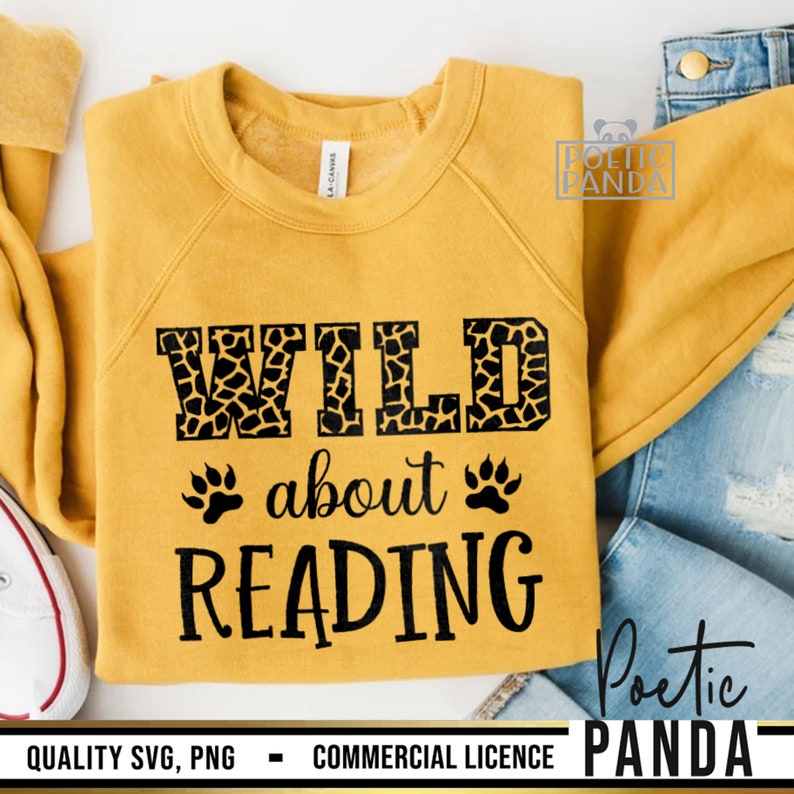 Puede incluir: Sudadera amarilla mostaza con el texto negro "WILD about READING". La palabra "WILD" tiene un estampado de jirafa. Huellas de patas a ambos lados de la palabra "about". La sudadera est&aacute; doblada sobre una superficie blanca con vaqueros.