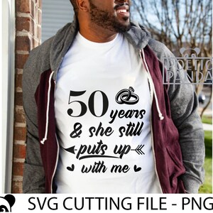May include: A white t-shirt with black text that reads "50 years & she still puts up with me" and two interlocking rings above the text. The text is centered on the shirt. The shirt is being worn by a person.