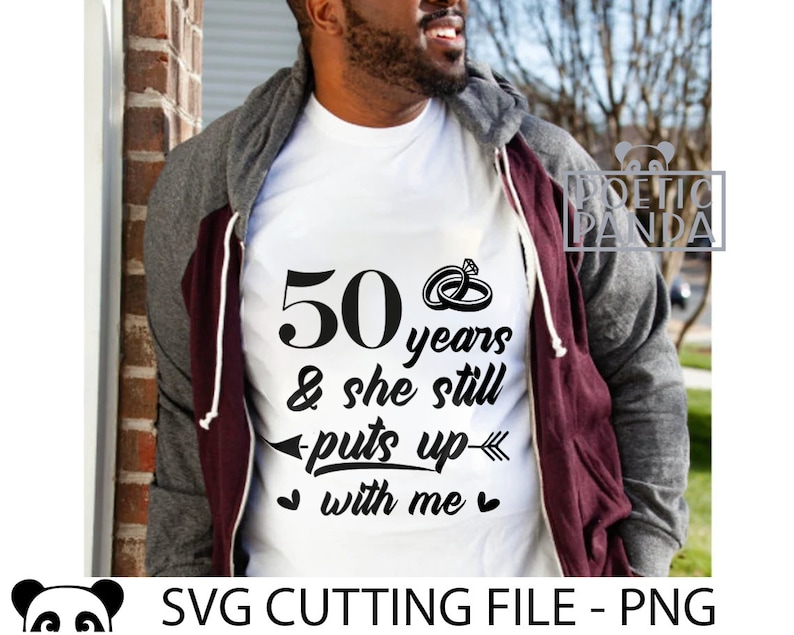 May include: A white t-shirt with black text that reads "50 years & she still puts up with me" and two interlocking rings above the text. The text is centered on the shirt. The shirt is being worn by a person.