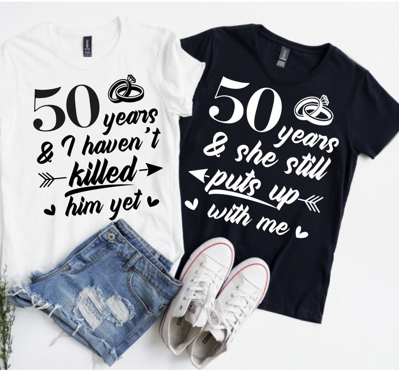 May include: Two white t-shirts with black text and graphics. The first shirt says "50 years & I haven't killed him yet" with an arrow pointing to the right. The second shirt says "50 years & she still puts up with me" with an arrow pointing to the left. Both shirts have a heart symbol on either side of the text.