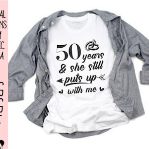 May include: White t-shirt with black text that reads "50 years & she still puts up with me" and a small drawing of two wedding rings.
