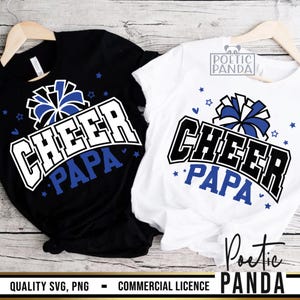 May include: Two t-shirts, one black and one white, each with the words "CHEER PAPA" in a bold, collegiate-style font. The design includes a blue and white pom-pom graphic and star accents. The shirts are displayed on wooden hangers.
