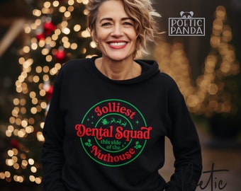 May include: A black hooded sweatshirt with the text "Jolliest Dental Squad this side of the Nuthouse" in red and green. The design includes a reindeer and stars. The sweatshirt is worn by a smiling person in front of a blurred Christmas tree.