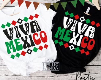May include: Two white and black t-shirts with the text "Viva Mexico" in green, red, and white. The text is in a bold, stylized font and is surrounded by a pattern of green, red, and white squares. The shirts are hanging on a wooden surface.