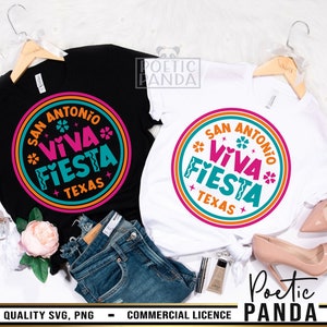 May include: Two white t-shirts with a black and white design featuring a circle with the text "San Antonio Viva Fiesta Texas" in pink, blue, and orange. The shirts are on a table with a pair of jeans, a makeup brush, and a bottle of makeup.