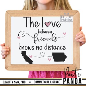 May include: A framed sign with the text "The love between friends knows no distance" with a pink heart and two black outlines of states connected by a dotted line with hearts. The sign is held by a person's hands.