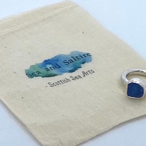 May include: A silver ring with a blue sea glass stone. The ring is in a small white drawstring bag with the text "Sea and Saltire - Scottish Sea Arts".