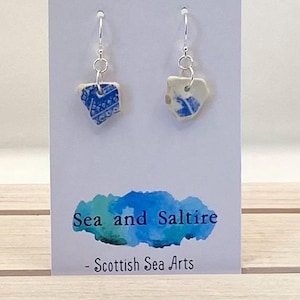 May include: Earrings with small, irregular ceramic pieces in blue and white patterns, hanging from silver hooks. The earrings are presented on a white card with the text "Sea and Saltire - Scottish Sea Arts" in blue, evoking a coastal theme.