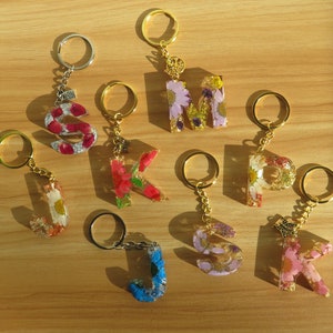 May include: A set of ten gold keychains featuring different letters of the alphabet. Each letter is crafted from clear resin, incorporating pressed flowers and leaves. These keychains are ideal for adding a touch of nature to your keys or bag.