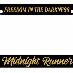 May include: Black license plate frame with gold text that reads "FREEDOM IN THE DARKNESS" at the top and "Midnight Runner" at the bottom.