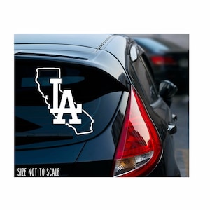 May include: White vinyl decal of the Los Angeles Dodgers logo, featuring the letters "LA" inside the outline of the state of California. The decal is on the back window of a black car. "Size not to scale" is printed below the decal.