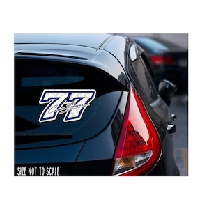 May include: A white and navy blue sticker with the number "77" and the name "Carson Hoover" on the rear window of a black car. The sticker has a bold, outlined design and is displayed on a car's rear window.