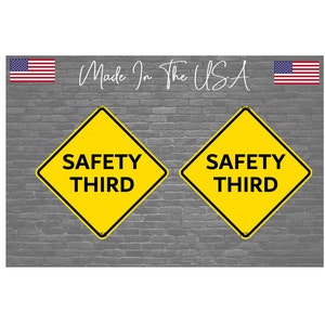 May include: Two yellow diamond-shaped road signs with black borders and black text that reads "SAFETY THIRD" on a gray brick wall background.