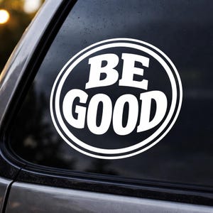 May include: White oval sticker with the words "BE GOOD" in bold, sans-serif font. The text is inside a white oval with a black border. The sticker is adhered to a car window.