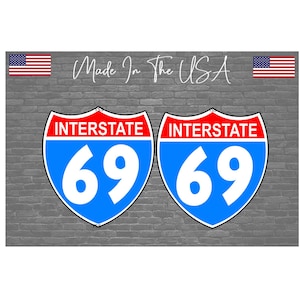 May include: Two blue and white Interstate 69 highway signs with red borders are displayed on a gray brick background. The words "Made in the USA" are written in cursive. American flags are at the top.