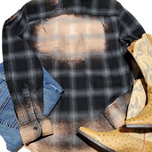 May include: A black and gray plaid flannel shirt with a bleached design on the back. The shirt is laid out on a white surface with a pair of brown cowboy boots and a pair of blue jeans.