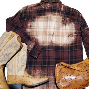 May include: A brown and black plaid flannel shirt with a bleached design. The shirt is paired with a brown leather handbag with metal studs and two brown leather cowboy boots.