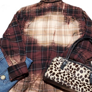 May include: A red and black plaid flannel shirt with a bleached design. The shirt is partially unbuttoned and has a leopard print handbag on top of it.