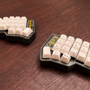 May include: Two split ergonomic keyboards with white keycaps and black frames are displayed on a brown surface. One keyboard has the text "FUCK" on the side, and the other has "THAT". The keyboards are designed for efficient typing.