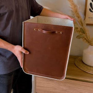 Puede incluir: Un contenedor de almacenamiento rectangular con un exterior de cuero liso de color marrón oscuro y un forro interior de color claro. El contenedor incluye un asa de cuero sujeta al panel frontal con remaches metálicos, lo que proporciona un diseño estructurado.