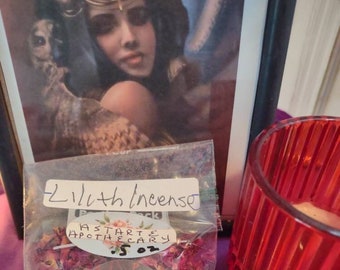 May include: A clear plastic bag labeled "Lilith Incense" filled with dried rose petals. A clear, round sticker with a red pentagram is on the bag. The bag is next to a red glass candle holder and a framed picture.