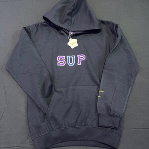 May include: Black hooded sweatshirt with the word "SUP" in purple and blue lettering across the chest. The sweatshirt has a front pocket and a tag that says "Tomi". The sleeve has text that says "est. 2020".