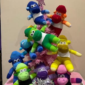 以下が含まれることがあります： ピンクのクリスマスツリーに、カラフルな靴下モンキーのオーナメントが飾られています。モンキーはそれぞれ異なる色のストライプを着ており、刺繍された顔をしています。ツリーの頂上にはピンクの星が飾られています。