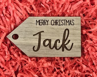 Könnte beinhalten: Ein Holz-Geschenkanhänger mit den Worten „MERRY CHRISTMAS“ und dem Namen „Jack“ in einer stilvollen Schrift graviert. Der Anhänger hat ein Loch zum Aufhängen und liegt auf einem Bett aus rotem Zerkleinerungspapier.