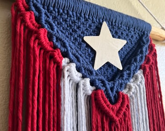 Puede incluir: Un tapiz de macramé con los colores de la bandera de Puerto Rico. La sección superior es azul marino con una estrella de madera, y la parte inferior presenta rayas verticales rojas y blancas. La pieza está suspendida de un palillo de madera.