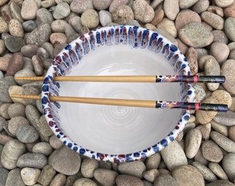 Puede incluir: Un cuenco de cerámica blanco con un borde decorativo con detalles azules y rojos. Dos palillos de madera con extremos estampados descansan sobre el cuenco. El cuenco está sobre un fondo de piedras de varios tamaños.
