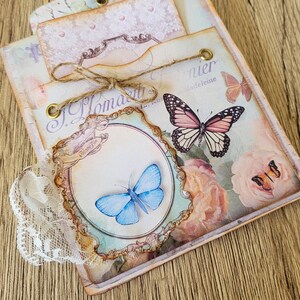 K&ouml;nnte beinhalten: Ein Scrapbook im Vintage-Stil mit floralem Design und Schmetterlingen. Das Scrapbook hat einen Spitzenbesatz und ein Etikett mit der Aufschrift "J. Homann" und "Madeleine".