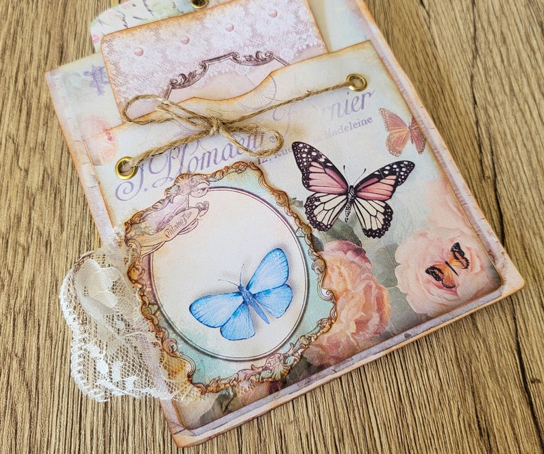 K&ouml;nnte beinhalten: Ein Scrapbook im Vintage-Stil mit floralem Design und Schmetterlingen. Das Scrapbook hat einen Spitzenbesatz und ein Etikett mit der Aufschrift "J. Homann" und "Madeleine".