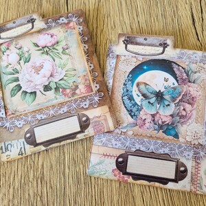 K&ouml;nnte beinhalten: Zwei dekorative Scrapbook-Papierumschl&auml;ge mit einer Spitzenbord&uuml;re. Ein Umschlag zeigt ein Blumenmuster mit rosa Pfingstrosen und einem Schmetterling. Der andere Umschlag zeigt einen Schmetterling mit blauen Fl&uuml;geln und einem Blumenmuster.