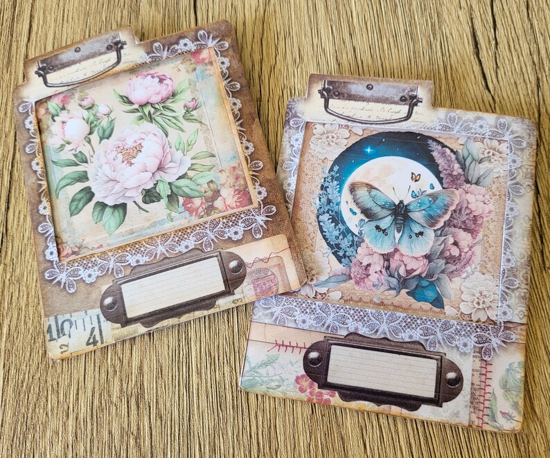 K&ouml;nnte beinhalten: Zwei dekorative Scrapbook-Papierumschl&auml;ge mit einer Spitzenbord&uuml;re. Ein Umschlag zeigt ein Blumenmuster mit rosa Pfingstrosen und einem Schmetterling. Der andere Umschlag zeigt einen Schmetterling mit blauen Fl&uuml;geln und einem Blumenmuster.