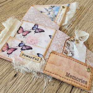 K&ouml;nnte beinhalten: Ein handgemachtes Scrapbook mit Vintage-&Auml;sthetik. Der Umschlag zeigt ein Blumenmuster mit Schmetterlingen und den Worten "be for you blossom every day". Die Innenseiten haben eine Vielzahl von dekorativen Elementen, darunter Spitze, Stoff und Papier. Das Scrapbook ist mit den Worten "Powerful" und "Memories" verziert.