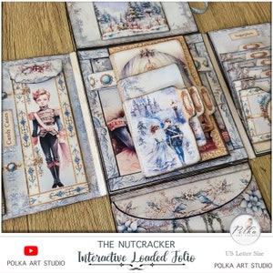Op de afbeelding: Een meerlaags papierkunstig folio met een winterthema. De voorkant heeft een blauwe en witte illustratie van een kasteel en een stel in winterkleding. De titel "The Nutcracker Interactive Loaded Folio" staat op de voorkant gedrukt. Het folio is ontworpen om te gebruiken als een scrapbook of dagboek.