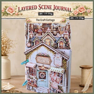 以下が含まれることがあります： The Craft Cottageというタイトルのレイヤードシーンジャーナル。Creative Atelierの看板、花のアクセント、青いドアの前に立つ人物が描かれた、多次元的な紙のイラストが特徴で、水色のバインディングリングが付いています。