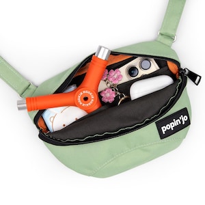 May include: A light green crossbody bag with a black zipper and strap. The bag is open, revealing an orange skate tool, a smartphone, wireless earbuds, and a small charm. The bag has the text "popin'jo" on the front.