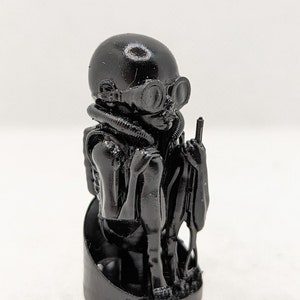 May include: A black figurine of a figure wearing a helmet, goggles, and a breathing apparatus. The figure is standing and has one arm raised. The figurine is on a round base.