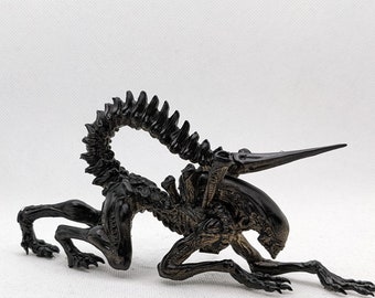 Puede incluir: Figurilla de criatura alienígena negra con una cola larga y una cabeza puntiaguda. La figurilla está en posición de cuclillas con las garras extendidas.