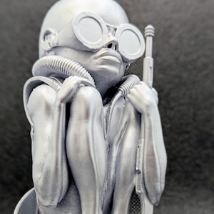 May include: A silver 3D printed figurine of a small, bald, humanoid figure wearing goggles and a breathing apparatus. The figure is sitting with its legs crossed and holding a rifle.