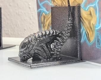 May include: A black and silver Alien-themed bookend featuring a detailed Xenomorph design. The bookend is made of a textured material and is designed to hold books upright. The background shows a skull illustration.