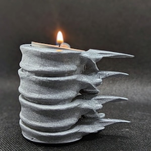 May include: A grey, textured candle holder with a jagged, organic design. A small tea light candle is burning inside the holder.