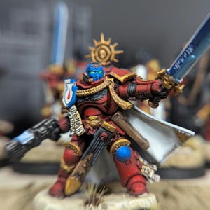 May include: A red and gold painted miniature figure of a space marine warrior holding a sword and a gun. The figure is wearing a white cape and has a blue face.